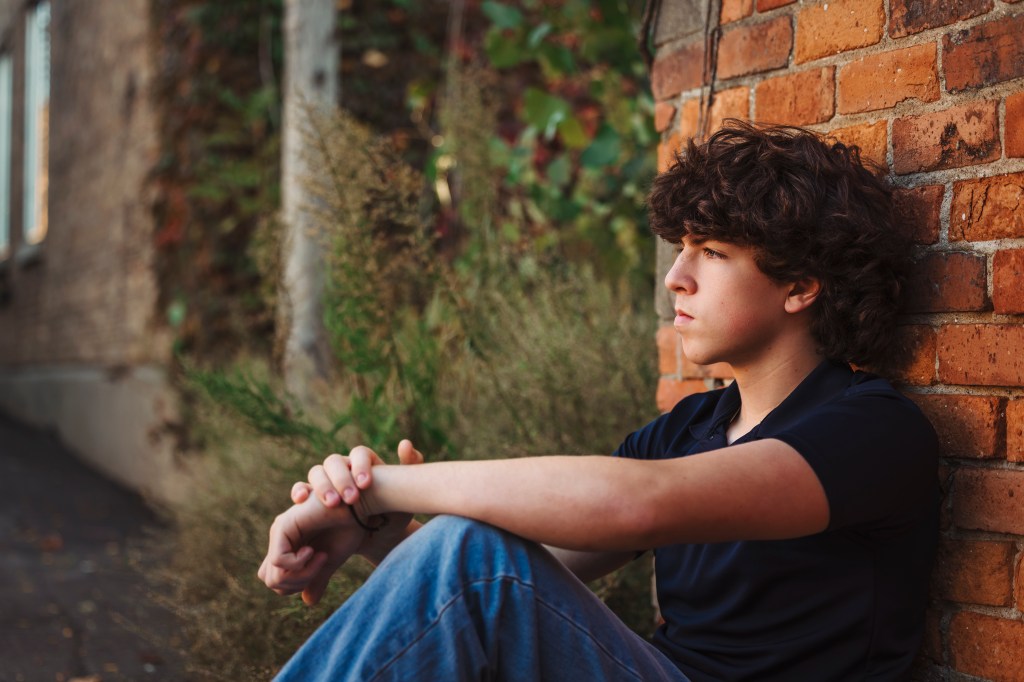 Senior boy sitting against a brick wall during an artistic senior photo session, using natural light and shallow depth of field