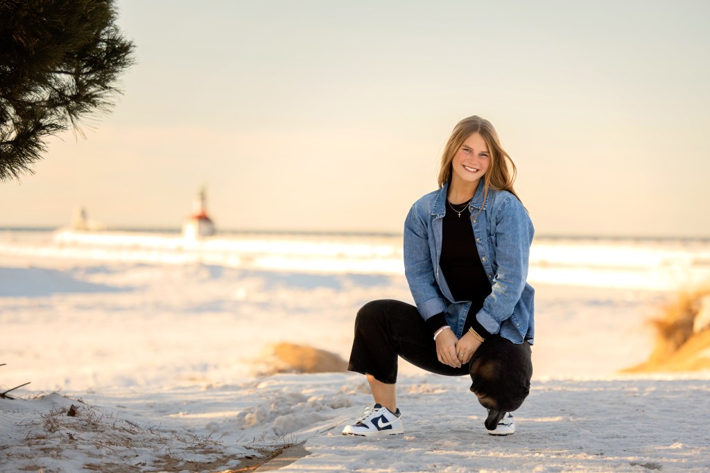 High school senior kneeling in the sand at Silver Beach in St. Joseph, Michigan with the lighthouse in the background on a sunny winter day.