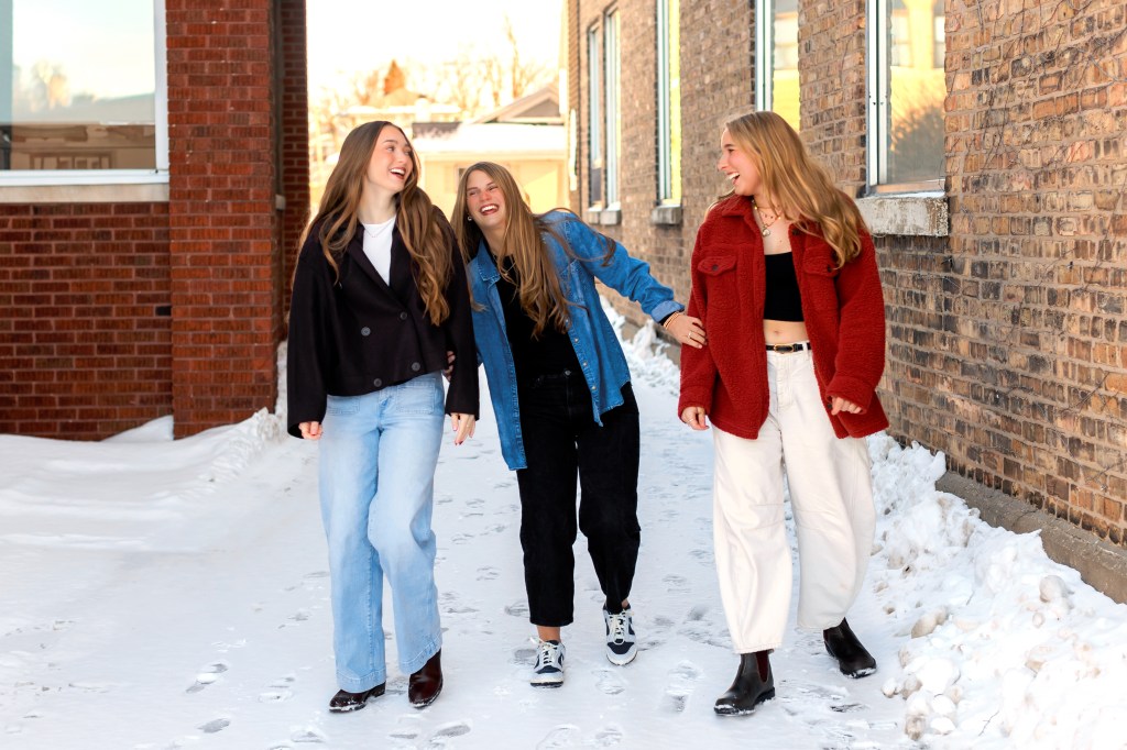 Three high school seniors walking together in downtown St. Joseph, Michigan on a bright winter day, photographed by a Southwest Michigan senior photographer with brick buildings and snow-lined sidewalks in the background.