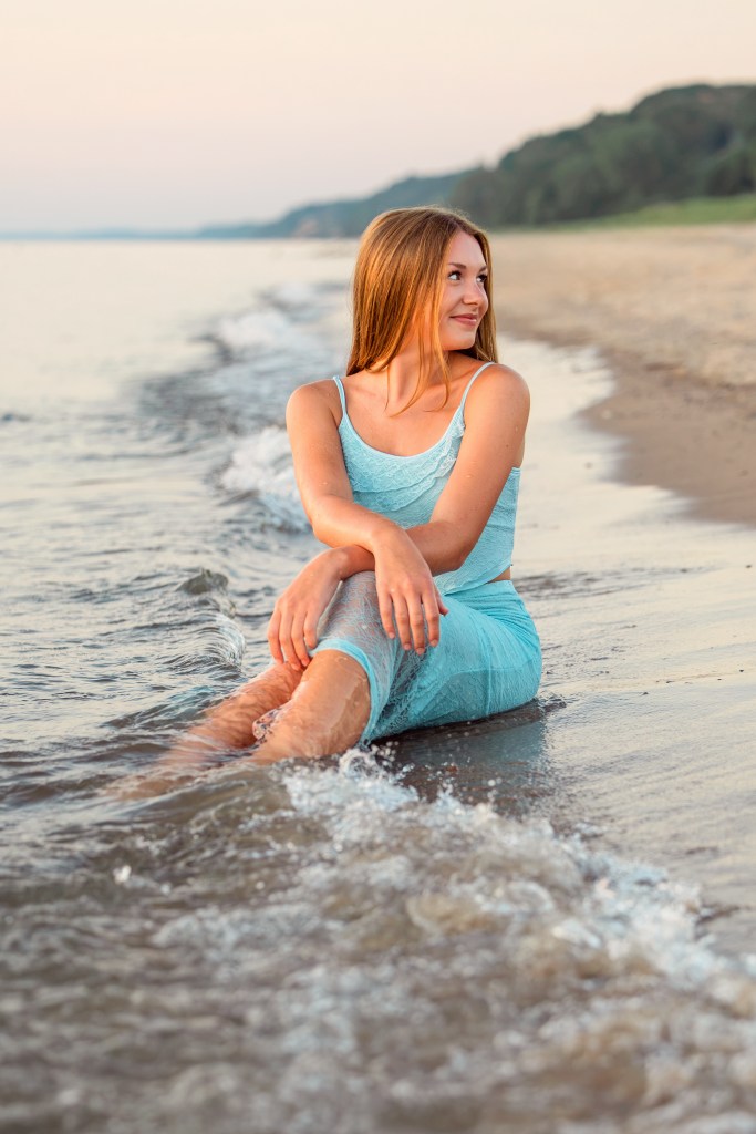 Emma smiling as gentle waves roll onto the shore at Jean Klock Beach during her Southwest Michigan senior portraits.