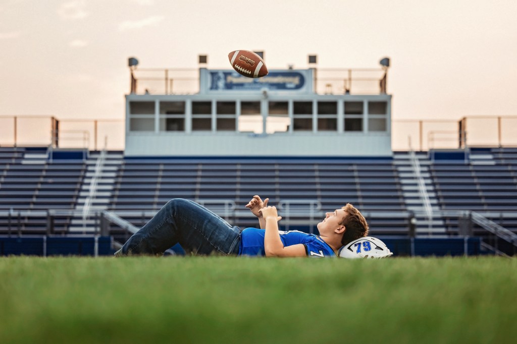 Lawton High School Class of 2027 senior lying on the football field and tossing a football in front of the Lawton Blue Devils stadium at sunset.
