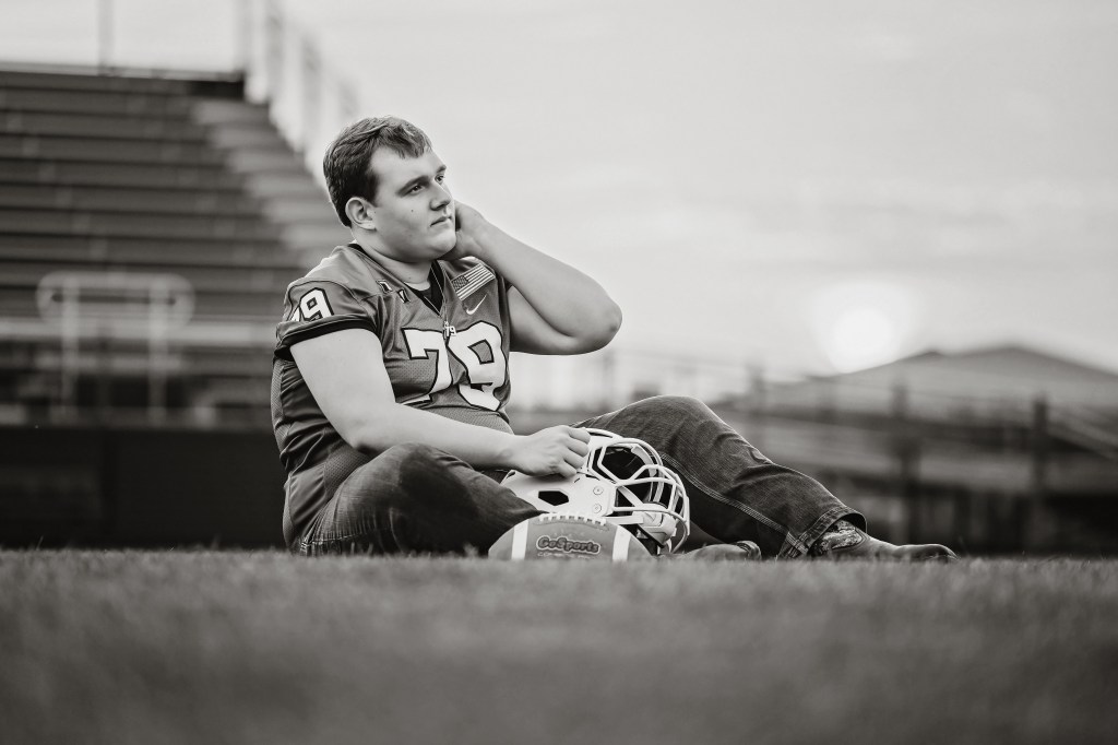 Black and white portrait of a Lawton High School Class of 2027 senior resting with his football helmet during senior pictures.