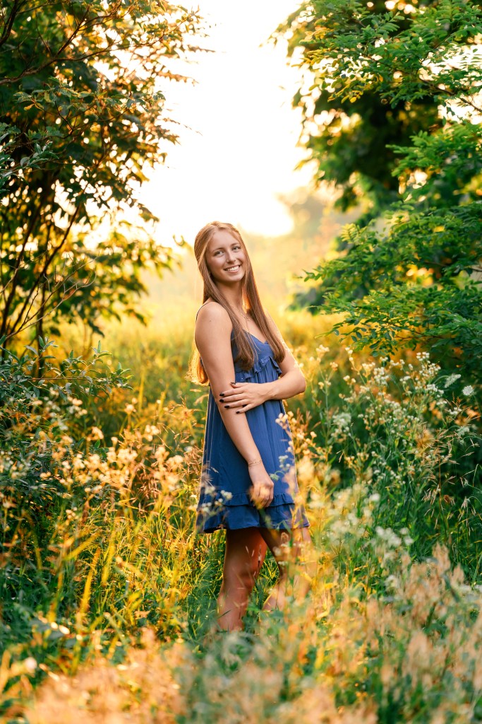 Class of 2027 female in blue dress posing in clearing with sunset in background